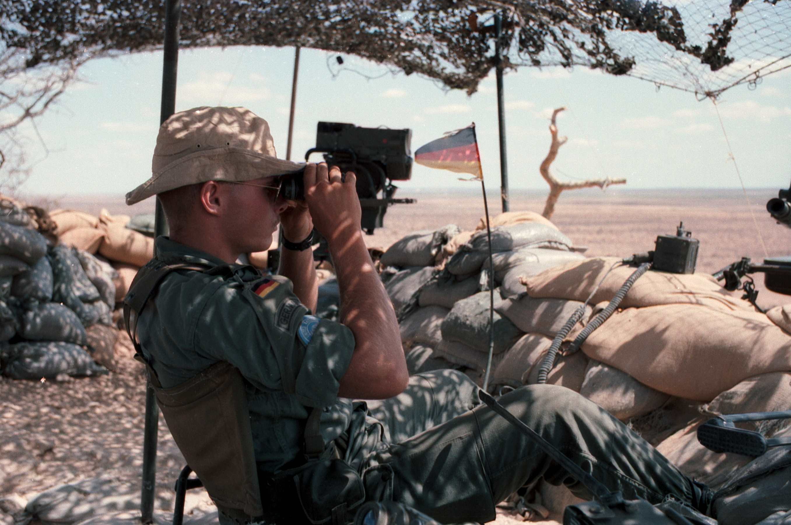 Auf einem Stuhl sitzender deutscher Soldat mit Fernglas und Laserentfernungsmesser. Im Hintergrund Sandwüste.
