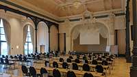 Großer Saal mit verzierten Säulen, Stuck an der Decke und mehreren Stuhlreihen zum Podest gewandt