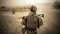 Ein Bundeswehrsoldat steht mit dem Rücken zur Kamera und sichert das staubige Gelände vor sich.