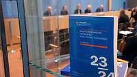 Aufgestelltes Cover des Berichts der Wirtschaftsweisen, die im Hintergrund bei einer Pressekonferenz zu sehen sind