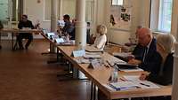 Menschen sitzen an Tischen in einem Raum mit Säulen und schauen zum Redner
