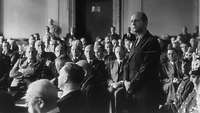 Ein Saal voller sitzender Menschen. In der Mitte steht ein Mann, die Hände überereinander gelegt.