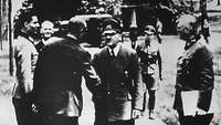 Drei Männer in militärischen Uniformen nehmen einen Mann in Uniform in Empfang.