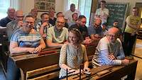 Menschen sitzen auf Schulbänken vor einer Schultafel