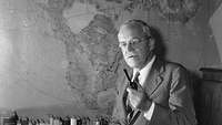 Portraitfoto des CIA-Direktors Allen W. Dulles mit Pfeife in der Hand vor einer Landkarte stehend.