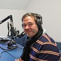 Drei Männer lächeln mit Kopfhörer vor dem Mikophon sitzend in die Kamera