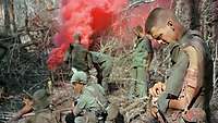 Mehrere Soldaten stehen im Dschungel, im Vordergrund ein verletzter Soldat, im Hintergrund Rauch
