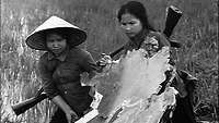 Zwei vietnamesische Frauen mit Gewehr auf dem Rücken tragen ein Trümmerteil durch ein Feld.