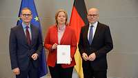 Zwei Männer und in der Mitte eine Frau stehen vor Flaggen, die Frau präsentiert ein Dokument.