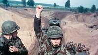 Zwei Soldaten auf einem Fahrzeug, einer hebt den Arm, dahinter weitere Fahrzeuge mit Soldaten
