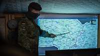 Ein Soldat steht vor einer Landkarte und deutet mit einem Zeigestock auf einen Punkt der Karte.