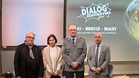 Drei Männer, darunter ein Offizier, und eine Frau stehen auf einem Podium vor einer Leinwand