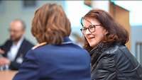 eine Frau in Uniform mit dem Rücken zur Kamera und eine Frau in dunkler Lederjacke mit Brille ist ihr Zugewand im Gespräch