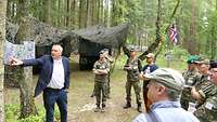 Ein Mann erklärt 8 Zuhörern in einem Waldstück alte Fotografien, im Hintergrund LKWs und Tarnnetze